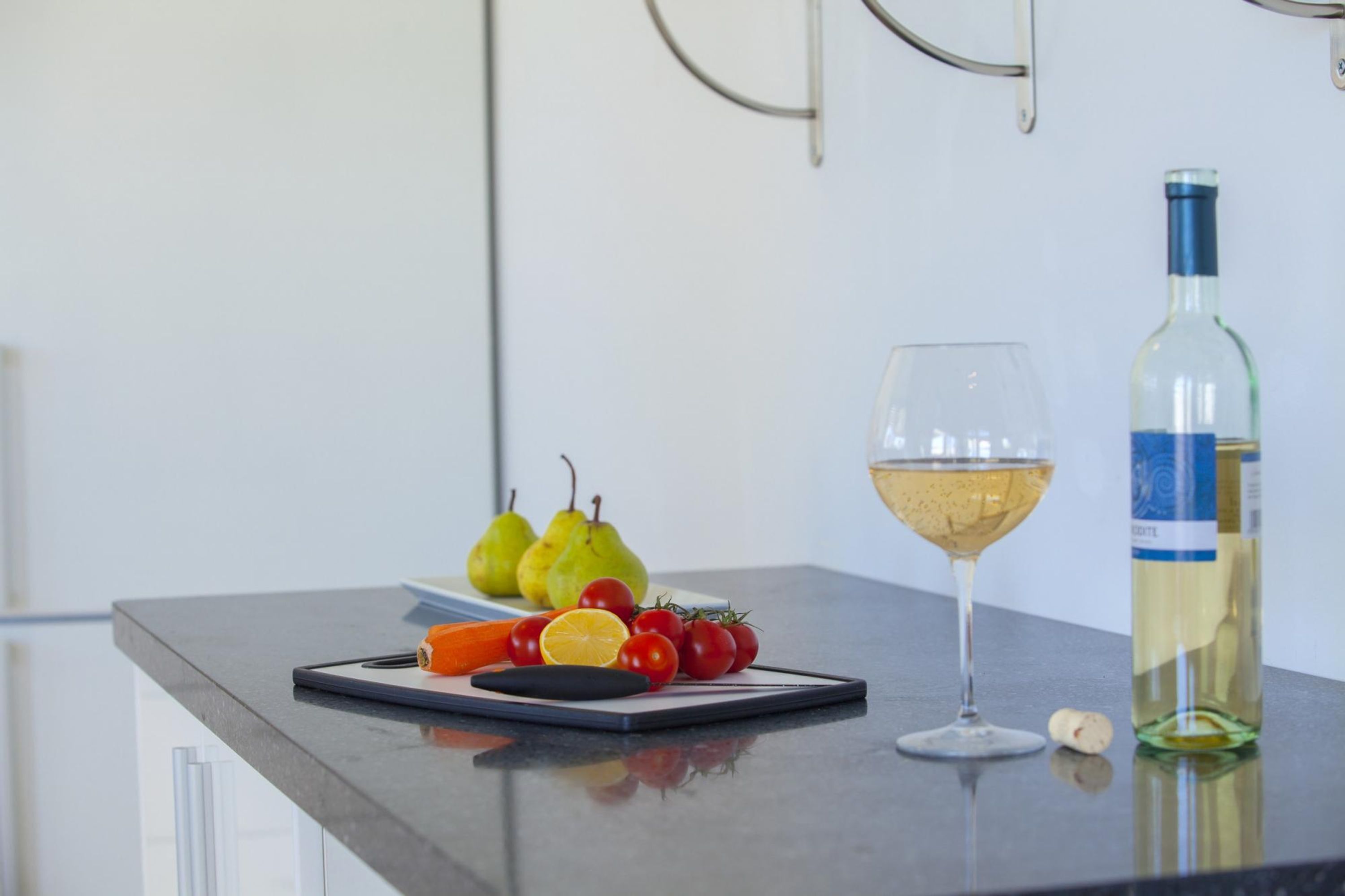 A kitchen counter with a wine glass, a bottle of white wine, and a plate with pears and tomatoes.