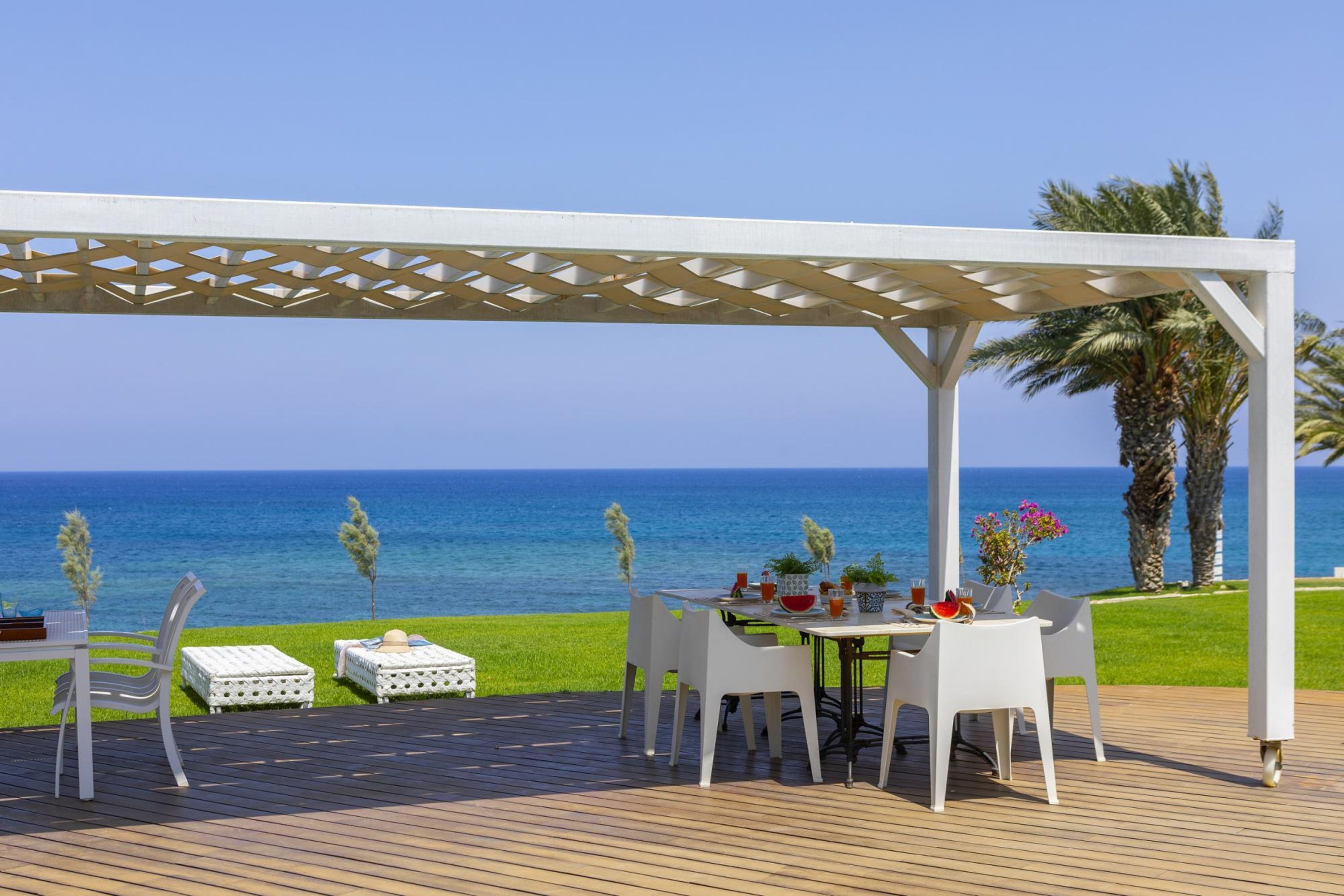A covered outdoor dining area with a view of the sea, white furniture, and a pergola structure.