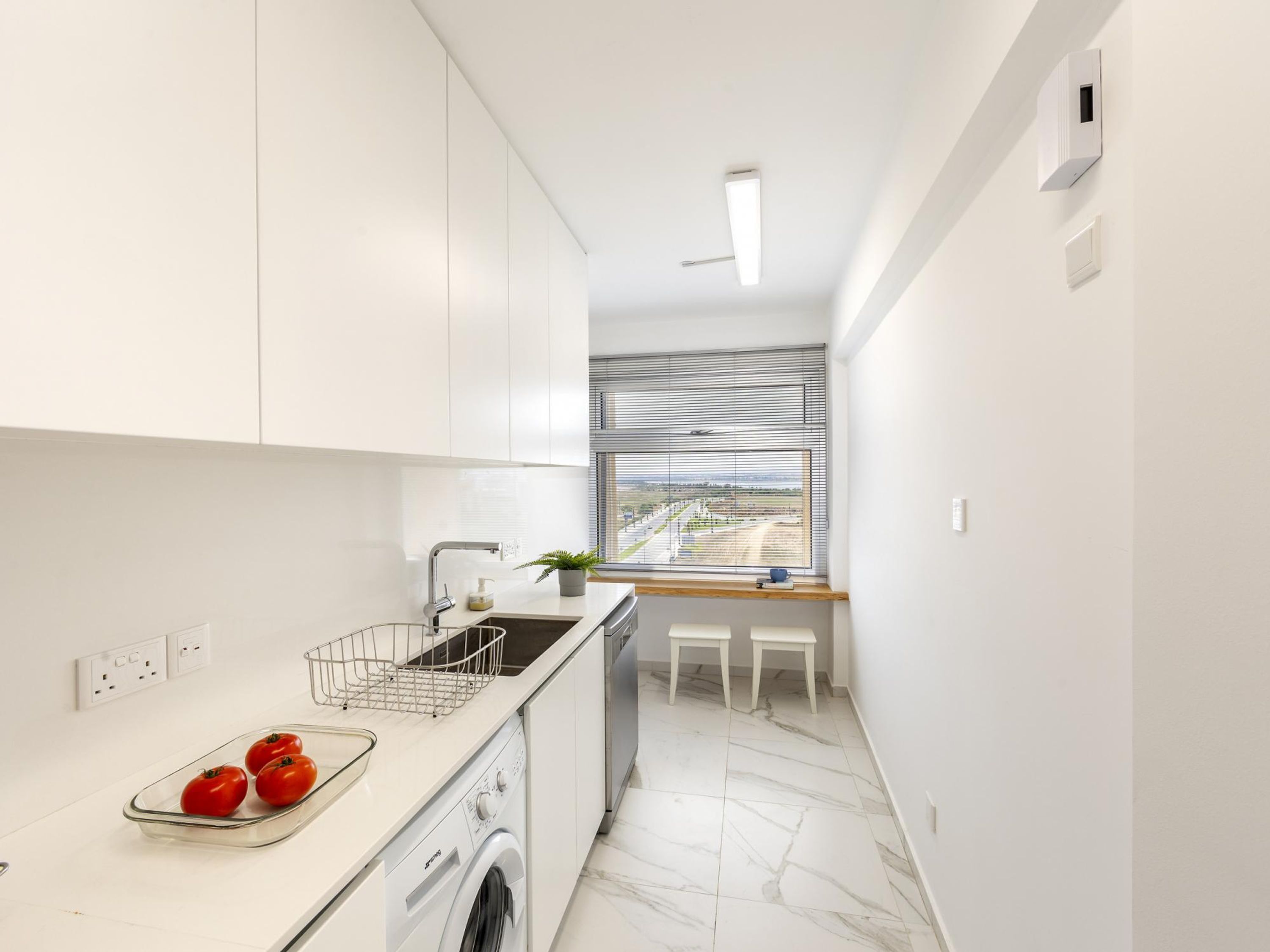 A modern kitchen with white cabinets, a built-in washing machine, a sink, and a window with blinds.