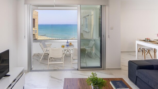 A modern apartment with a balcony overlooking the sea, a dining area, and a living room with a sofa and a TV.