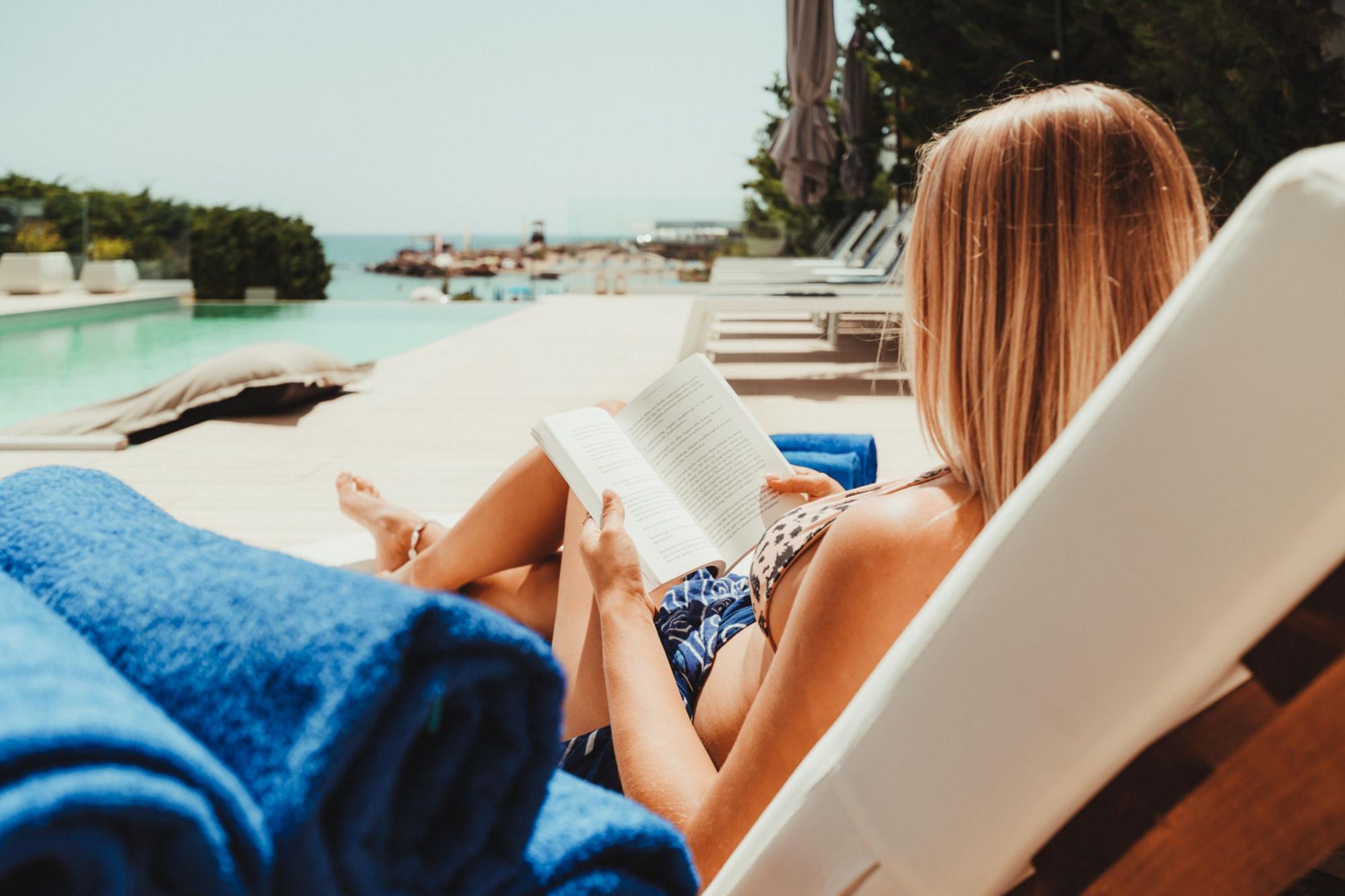 Eine Person entspannt auf einer Liege am Pool mit einem Buch, mit Blick auf das klare Meer.