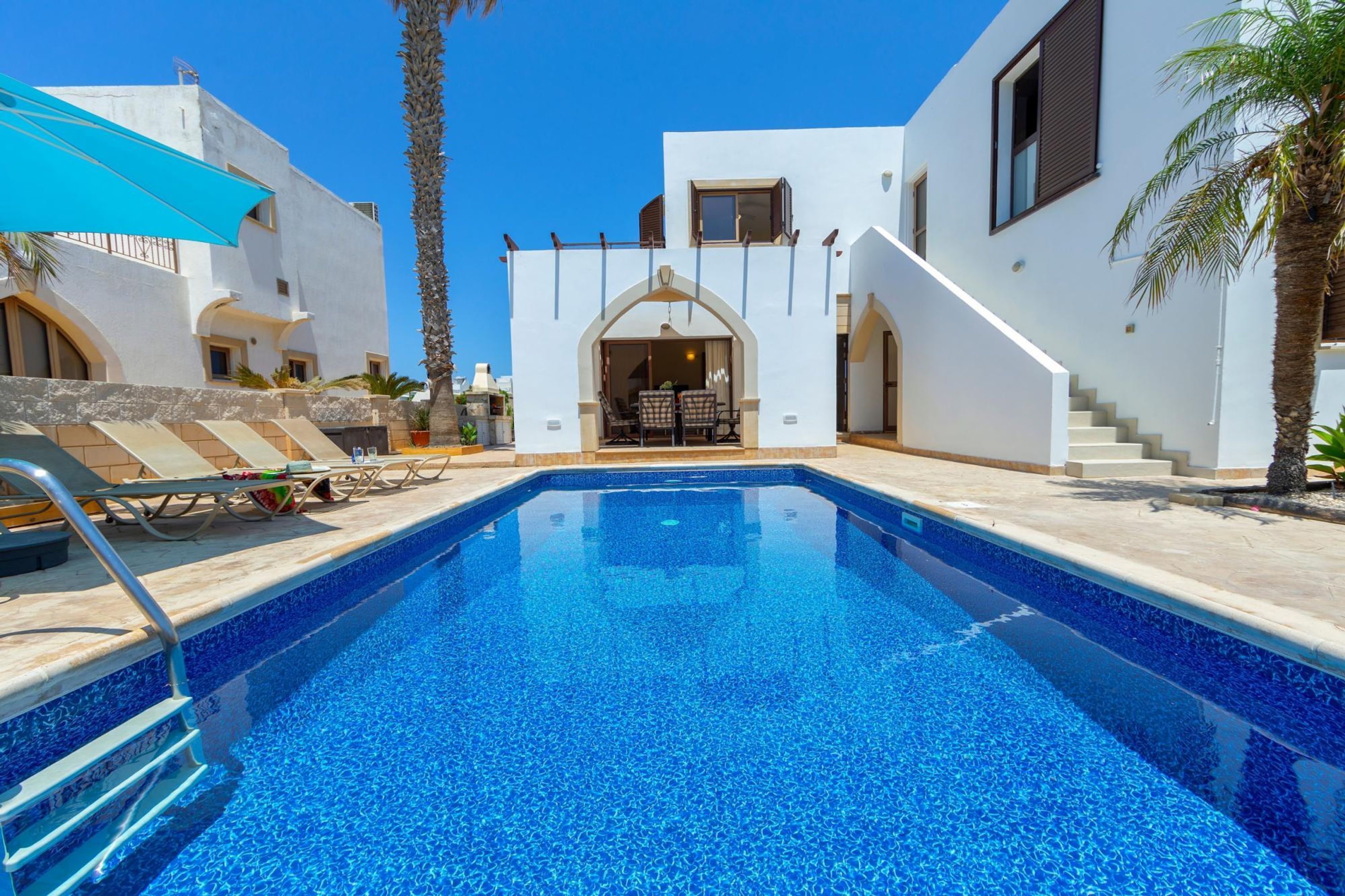 A swimming pool with a clear blue water, surrounded by white buildings with arched doorways and a staircase leading to the upper floor.