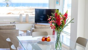 A modern living room with a glass table, a vase of flowers, and a plate of fruit. A television is mounted on the wall, and there is a view of the beach through large windows. - Protaras Seafront Villa Violetta (Paralimni)