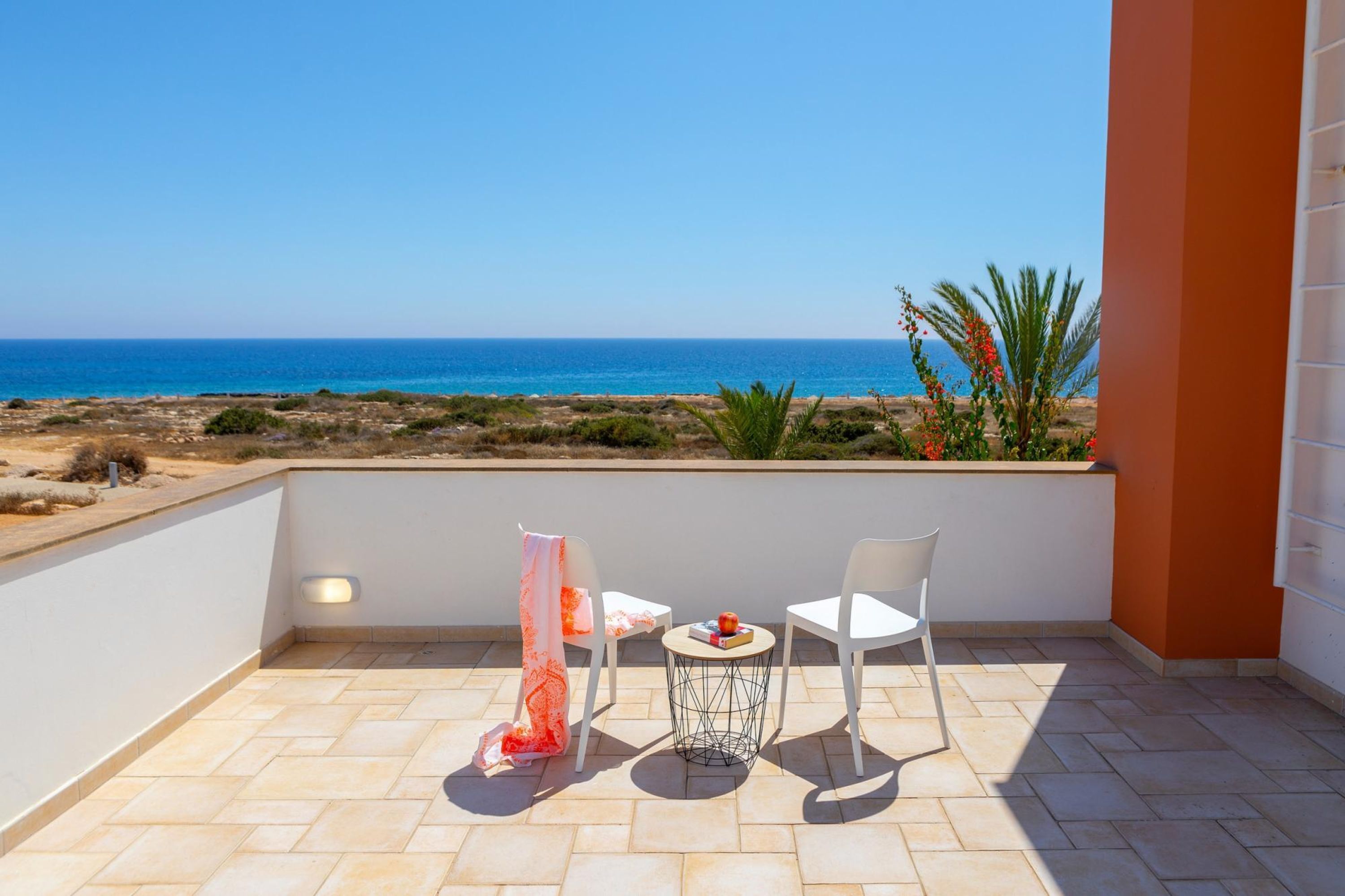 A balcony with a view of the sea, two white chairs, a small round table with a book and fruit, and a decorative cloth.