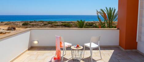 Un balcón con vista al mar, dos sillas blancas, una mesita redonda con un libro y fruta, y un textil decorativo.