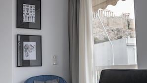 A room with a view of ancient ruins through a window, a framed picture of classical architecture, and a blue chair with a grey pillow. - Deco Marble Apt With Acropolis View (Athens)