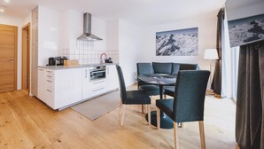 Een moderne keuken met witte kastjes, een ingebouwde oven en een roestvrijstalen afzuigkap. Er is een eethoek met een tafel en stoelen, en een groot raam met gordijnen.