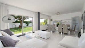 Ein modernes Wohnzimmer mit weißen Möbeln, einem Essbereich mit gemusterten Stühlen und großen Fenstern mit Blick ins Freie.