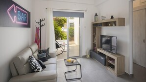 Una sala de estar moderna con un sofá, una mesa de centro, un televisor y una estantería. Una puerta da acceso a un espacio exterior con plantas.