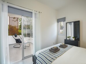 A modern bedroom with a large bed, a sliding glass door leading to an outdoor seating area, a dark dresser, and a mirror. - Ayios Elias Alice Suite (Paralimni)