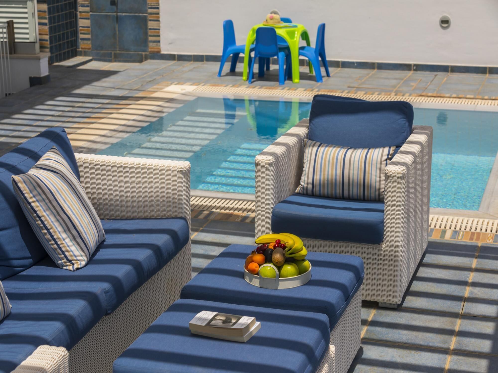 A poolside area with two wicker sofas, a table with chairs, and a fruit bowl on a table.