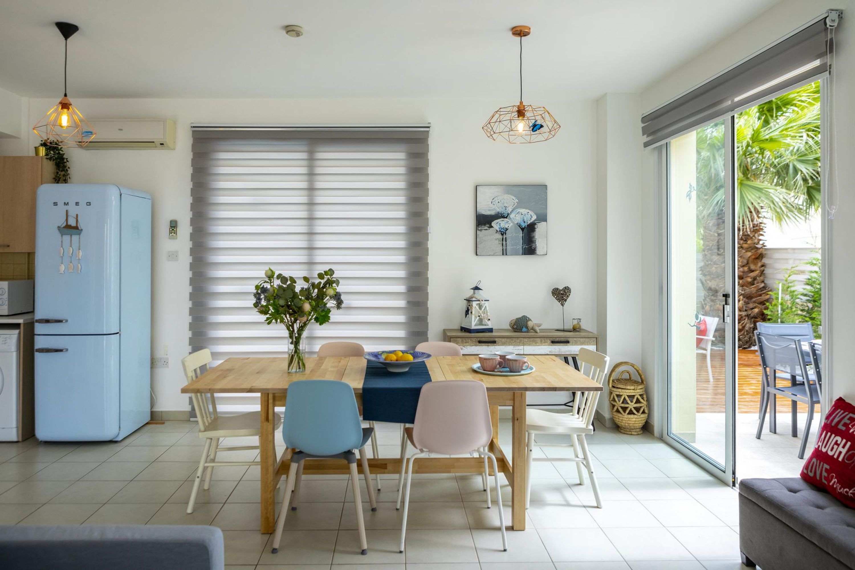 A modern kitchen with a blue refrigerator, a dining table with chairs, and a large window with blinds.