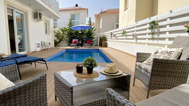 A poolside area with wicker furniture, a table with a hat, and a blue umbrella. - Villa Kalimera in Protaras (Paralimni)