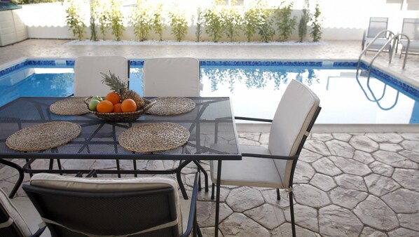 A poolside table with woven placemats, a fruit bowl, and white chairs.