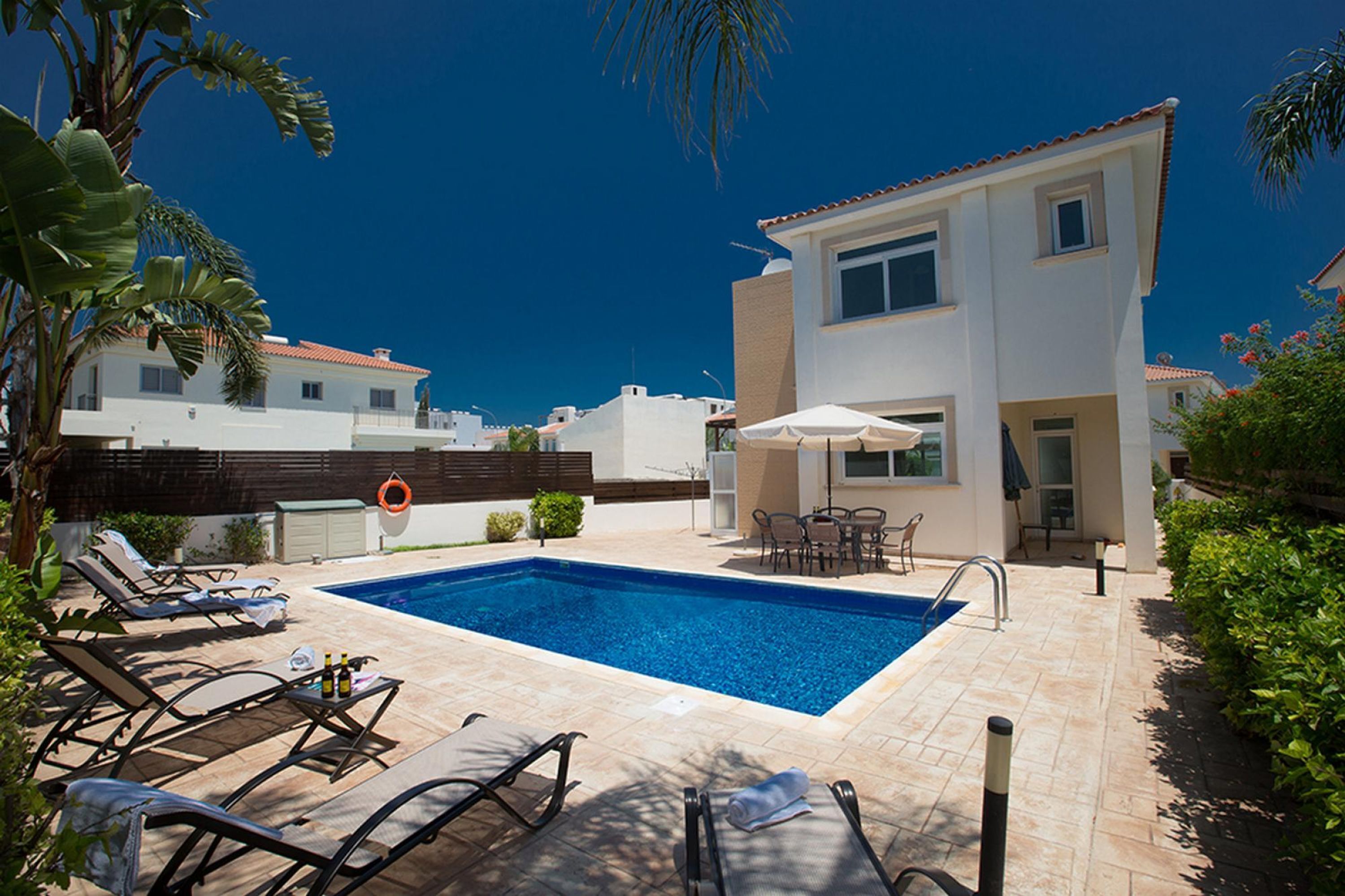 A pool area with lounge chairs and a table, surrounded by a white building with a tiled roof.