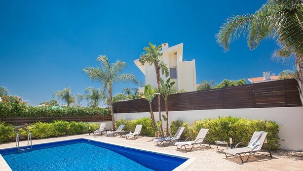 Una piscina con sillones de descanso, palmeras y un edificio moderno al fondo.