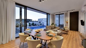 Una sala moderna con una mesa de comedor de madera, sillas color beige, un sofá y un ventanal con vista a una piscina y un área exterior.
