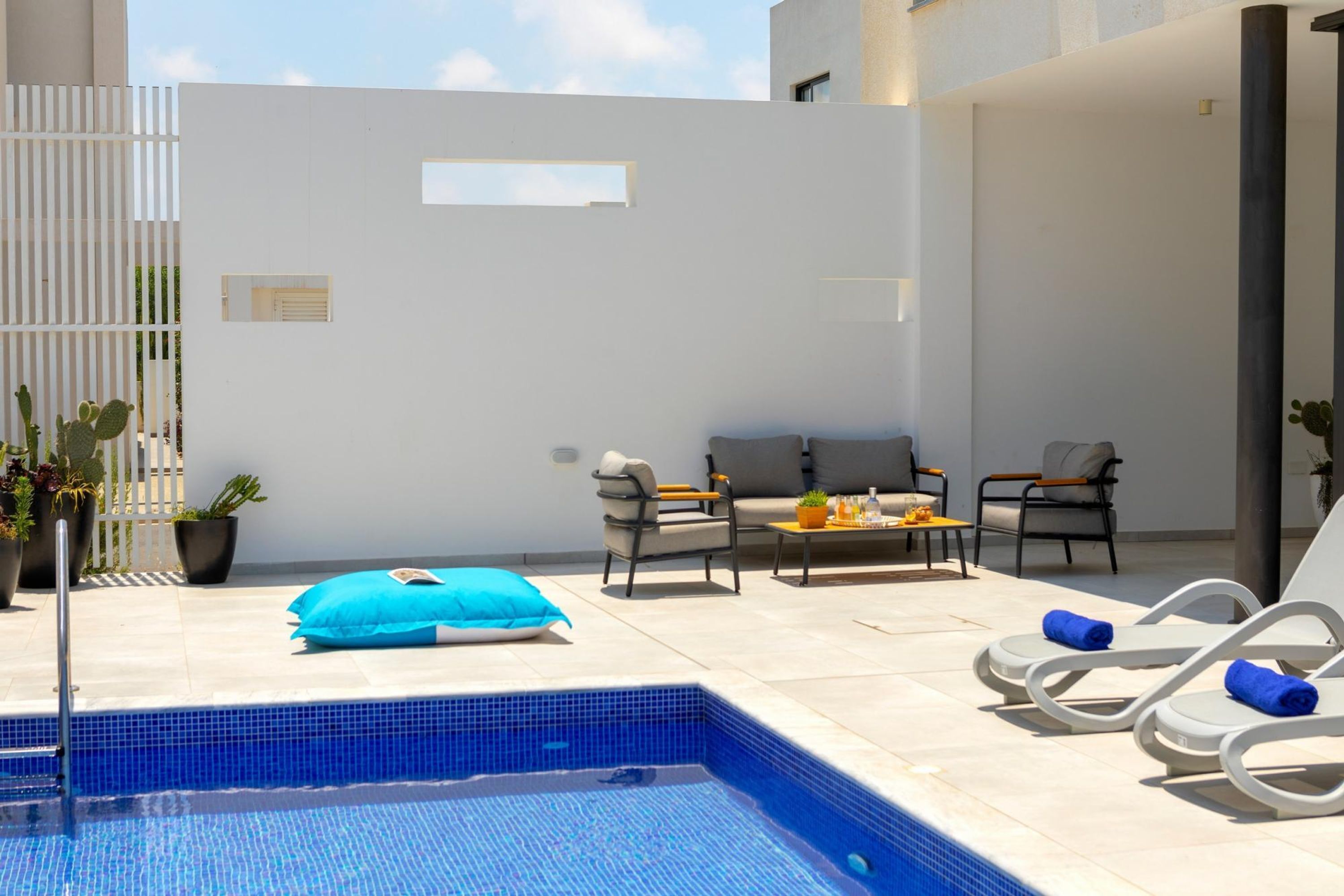 A modern outdoor pool area with a blue-tiled pool, lounge chairs, a sofa, and potted plants.