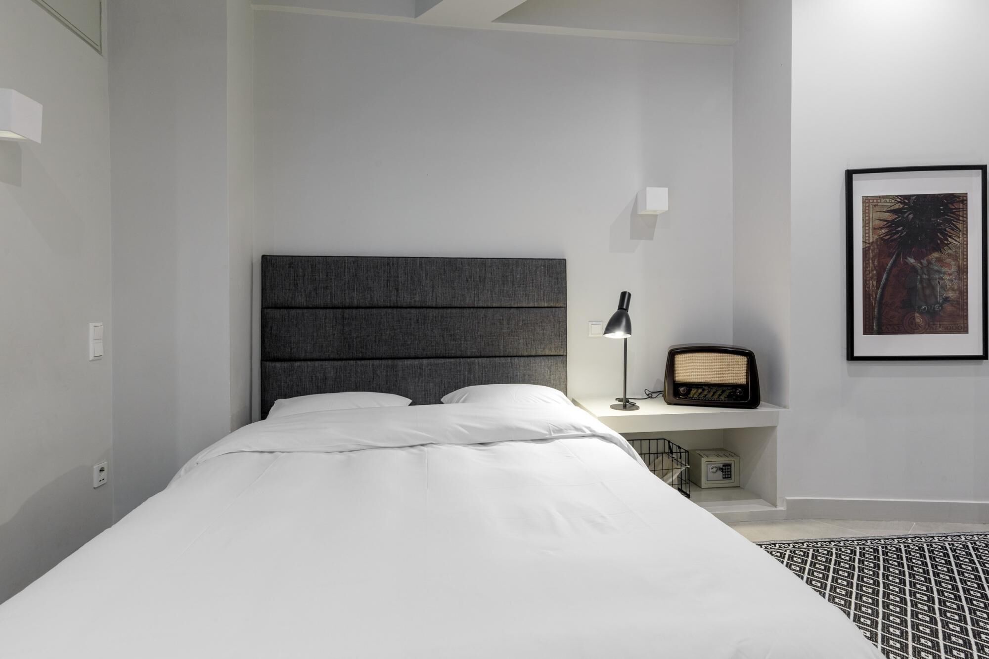 A modern bedroom with a large bed, a bedside lamp, a radio, and a framed picture on the wall.
