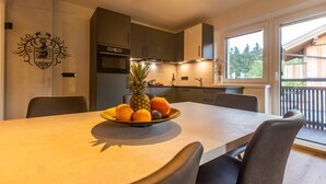 Une cuisine moderne avec une table à manger pour quatre personnes, agrémentée d’un panier de fruits contenant un ananas, des oranges et un avocat.
