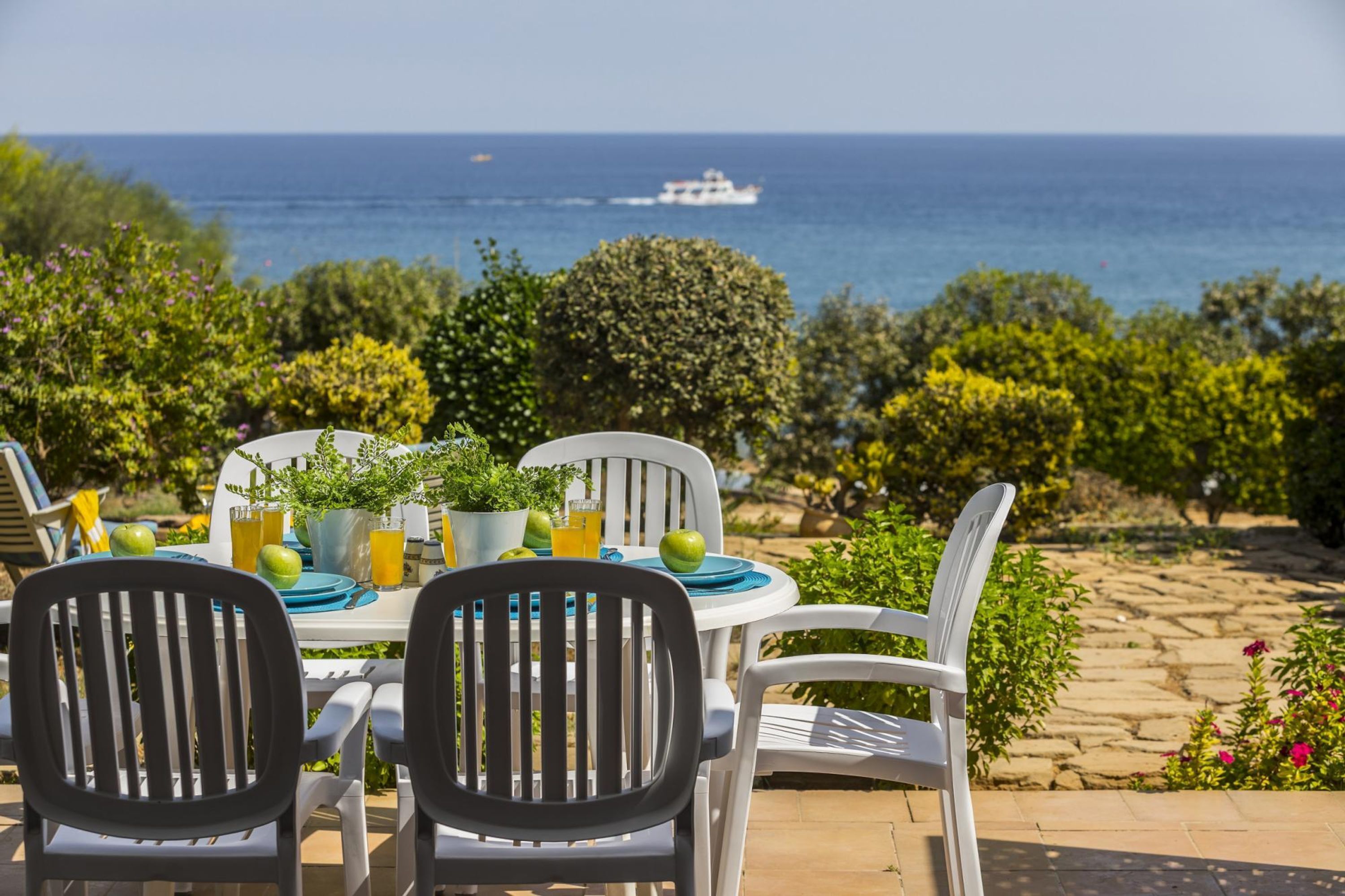 An outdoor dining area with a table set for two, surrounded by white chairs, overlooking a scenic ocean view with a boat in the distance.