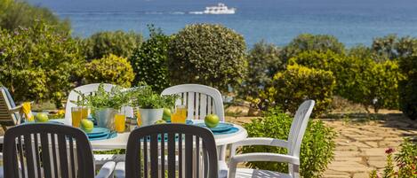 An outdoor dining area with a table set for two, surrounded by white chairs, overlooking a scenic ocean view with a boat in the distance.