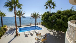 A pool area with a view of the sea, surrounded by palm trees and a dining setup. - Sunset Beachfront Villa Meneou (Larnaca)