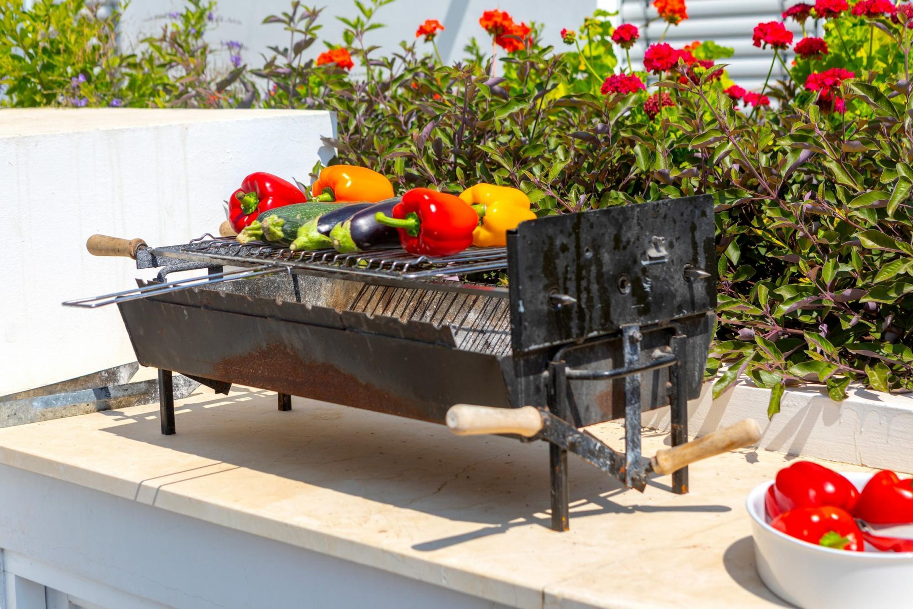 Una parrilla portátil con verduras encima, ubicada en un balcón con barandilla blanca y un jardín de flores al fondo.