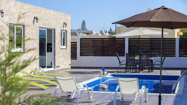 Un área de piscina moderna al aire libre con sillones de descanso, una mesa con sillas y un gran paraguas.