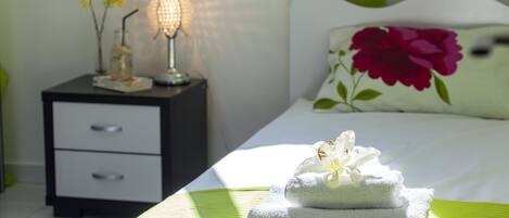 A neatly made bed with a floral pillow, a bedside lamp, and a stack of towels on a green bedspread.