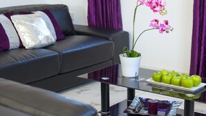 Una sala moderna con un sofá de cuero negro, una mesa de centro de vidrio, un jarrón blanco con una orquídea rosa y una bandeja de manzanas verdes.