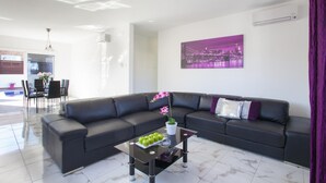 Una sala moderna con un sofá de cuero negro, una mesa de centro de vidrio con manzanas verdes, y un cuadro de un paisaje urbano morado en la pared.