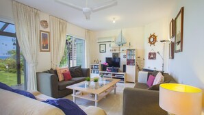 A living room with a sofa, coffee table, and TV. There is a kitchen area with a TV and a dining table. The room has large windows and curtains. - Protaras Coast Panorama (Paralimni)