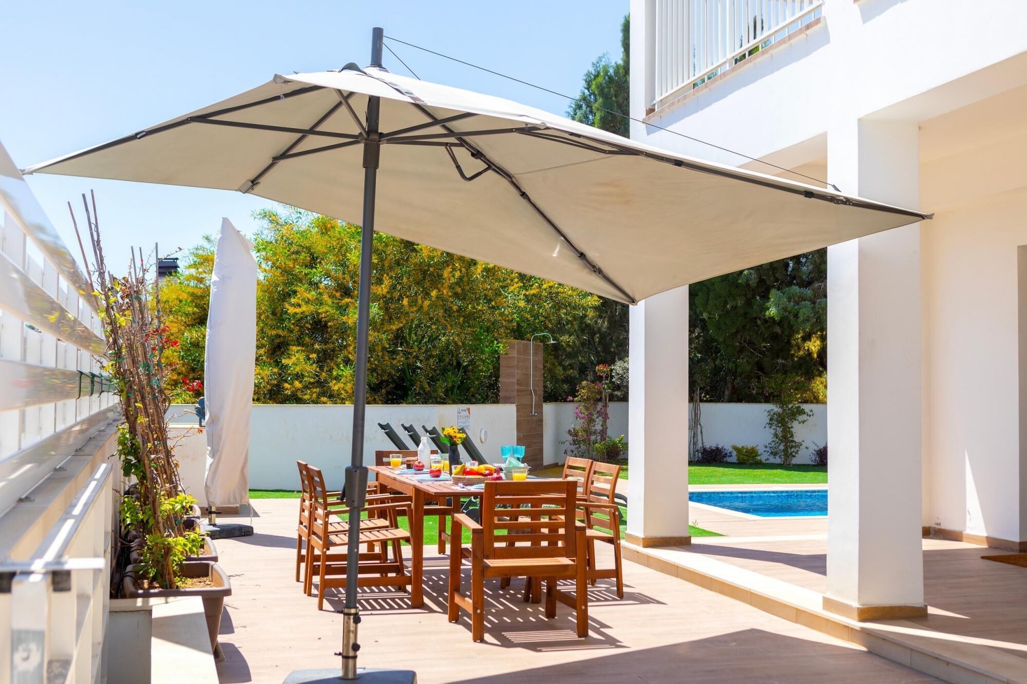 An outdoor patio with a table and chairs, a large umbrella, and a pool in the background.