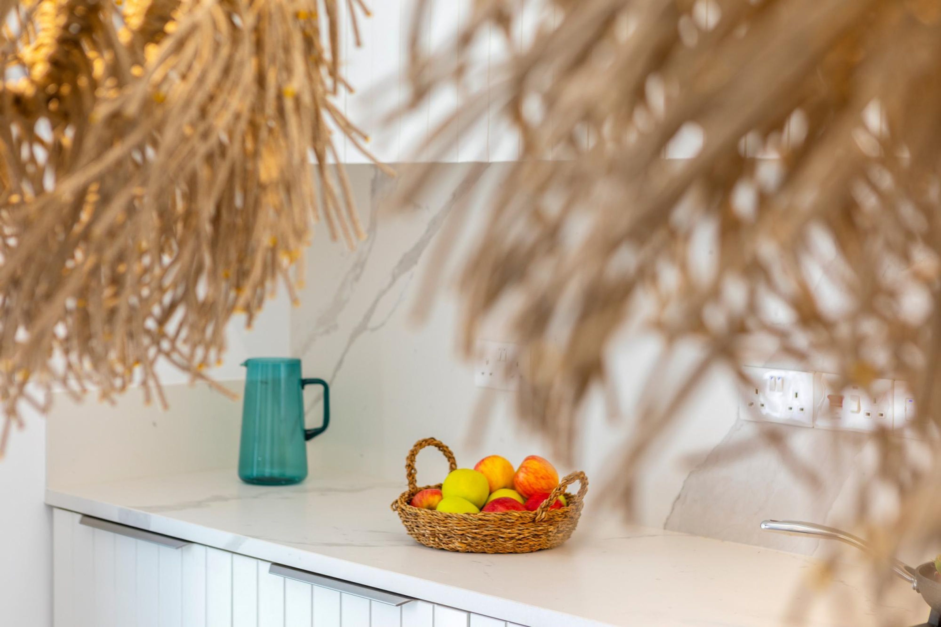 A kitchen counter with a wicker basket containing fruits, a teal pitcher, and a white cabinet.