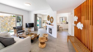 A modern living room with a sofa, coffee table, and wooden stools. There is a TV, a fireplace, and a kitchen area in the background. - Protaras Rose By The Sea Villa (Paralimni)