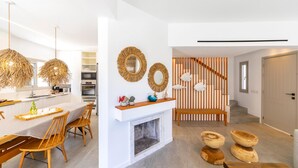 A modern kitchen with white cabinetry, a dining area with wooden chairs, a fireplace, and a staircase leading to a loft bedroom. - Protaras Rose By The Sea Villa (Paralimni)
