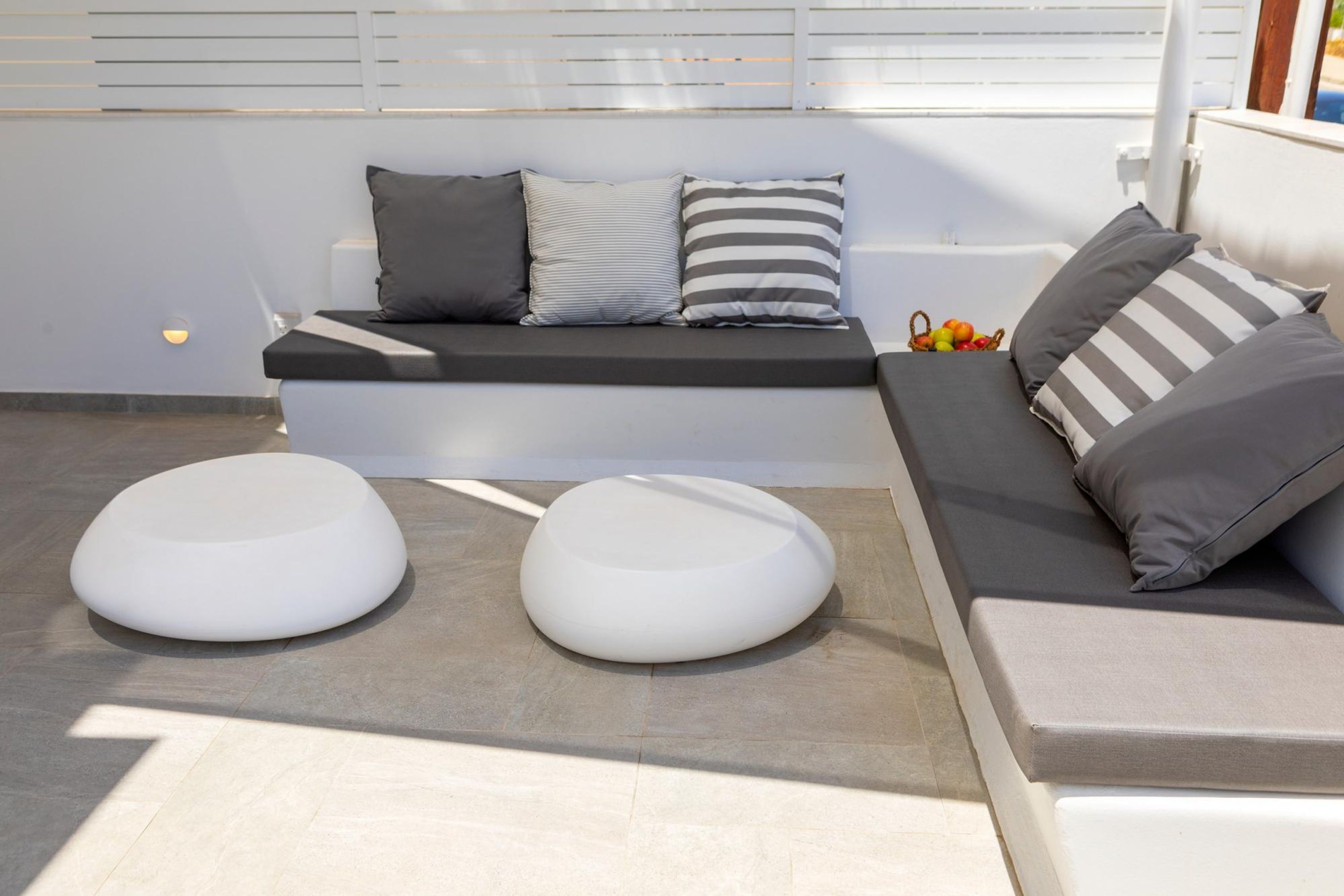A modern outdoor seating area with a bench and two round white ottomans, a white wall with horizontal blinds, and a window with a view.