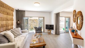 A modern living room with a sofa, coffee table, and TV. There are curtains, a stone wall, and a view of the outdoors. - Protaras Rose By The Sea Villa (Paralimni)