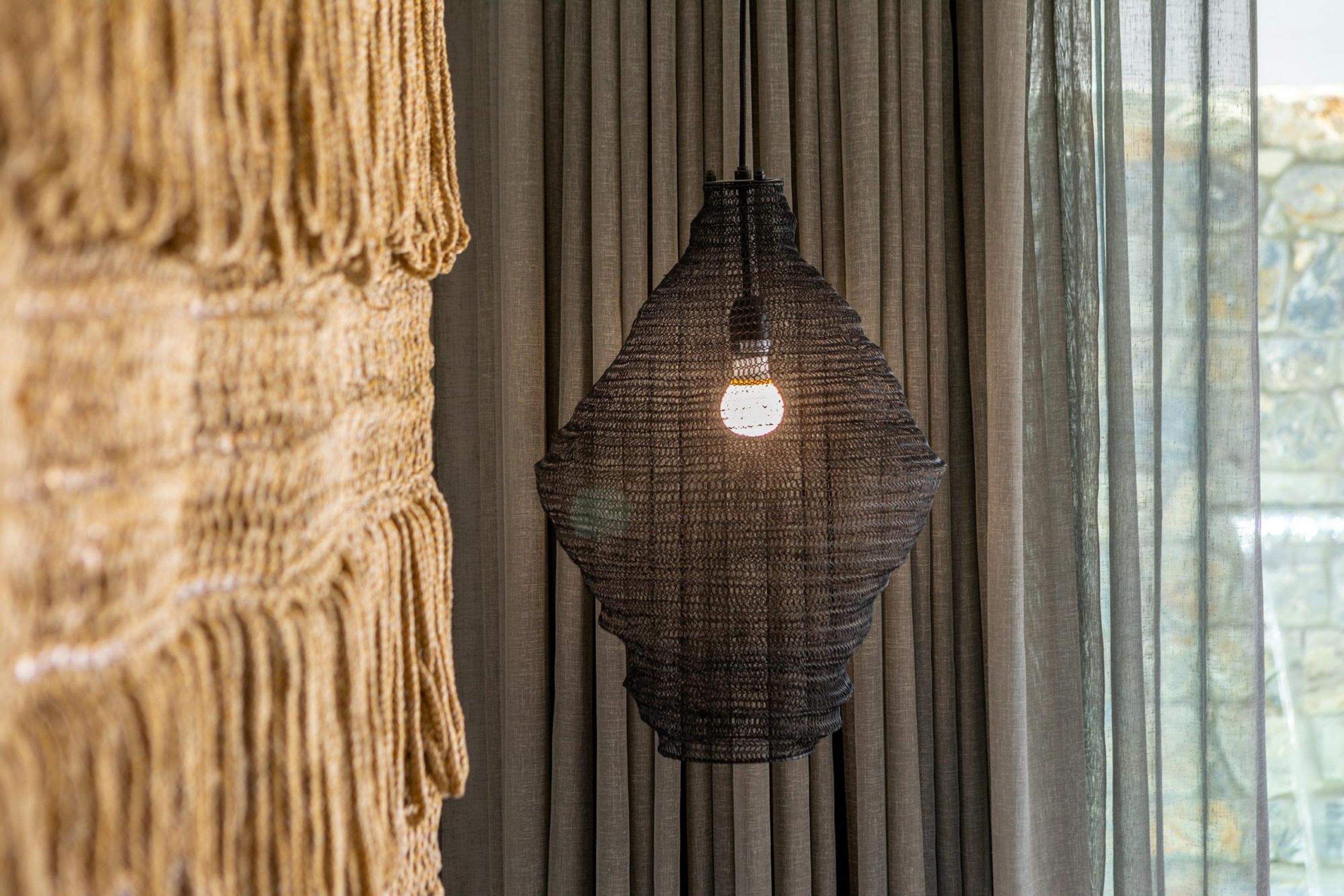 Lámpara colgante con estructura tejida y texturizada, ubicada frente a una ventana con persianas verticales.