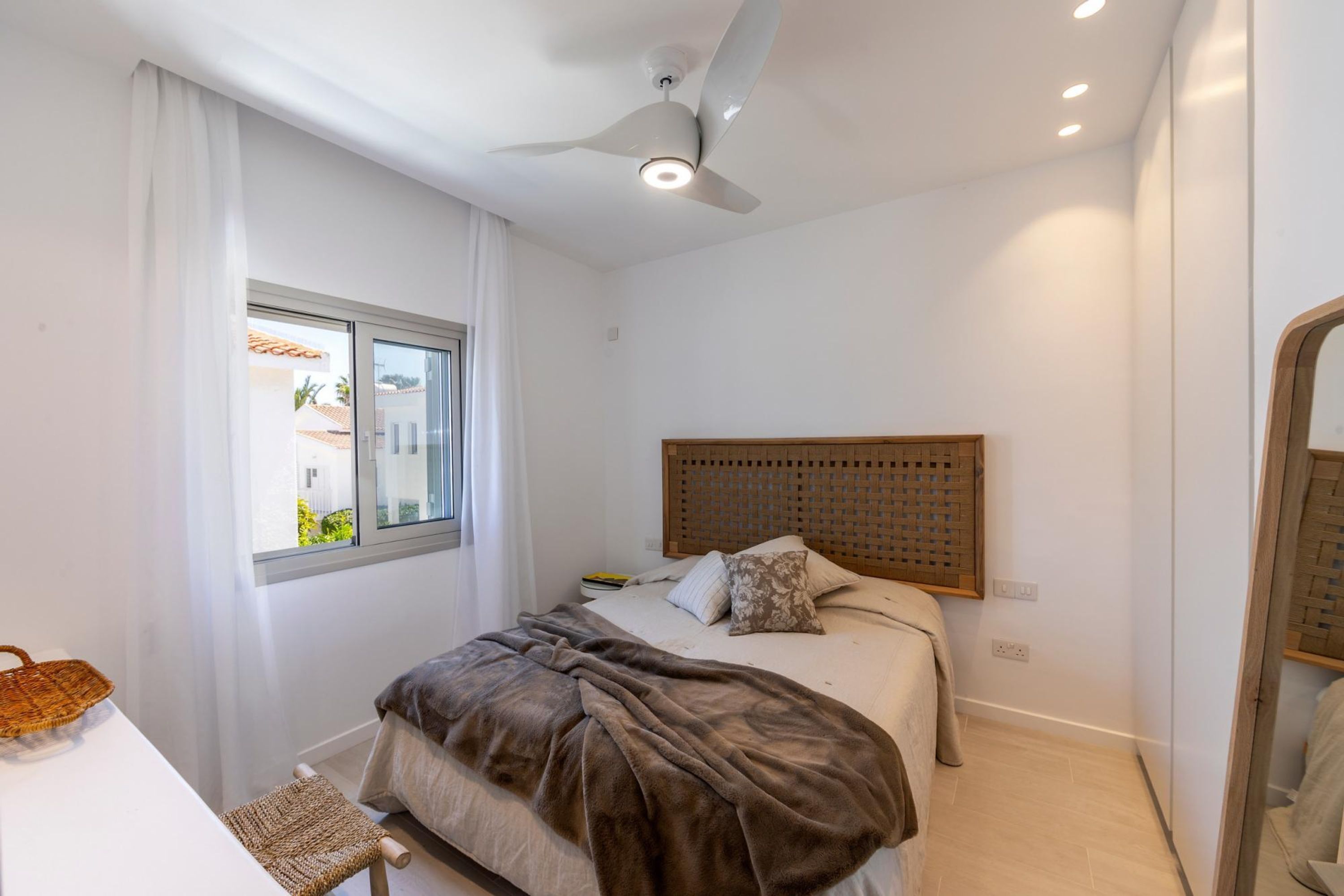 Ein Schlafzimmer mit einem Bett, Deckenventilator, einem Fenster mit Vorhängen, einem Nachttisch und einem gewobenen Stuhl.