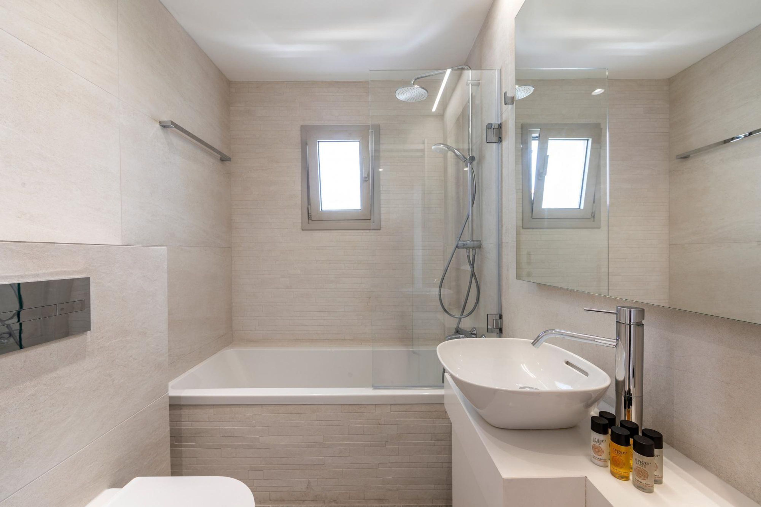 A modern bathroom with a bathtub, a sink, and a shower. There are two small windows and a large mirror.