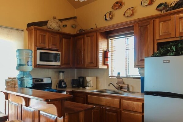 Una cocina con armarios de madera, microondas, refrigerador, dispensador de agua y una ventana con persianas.