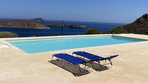 Una piscina rectangular con agua azul cristalina, rodeada de baldosas color beige. Hay tres sillas de descanso azules con estructura metálica y cojines negros. La piscina está ubicada frente a un terreno rocoso y el mar.