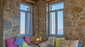 Una habitación acogedora con paredes de piedra, vigas de madera en el techo y un ventanal grande. Hay un sofá blanco con cojines de colores y una mesa de centro de madera.