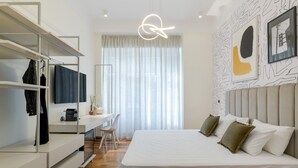 Un dormitorio moderno con cama, escritorio y silla. Se observa un armario con ropa colgada y un perchero.