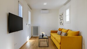 Una sala de estar con un sofá amarillo, una mesa de centro negra, un televisor de pantalla plana montado en la pared y un radiador.
