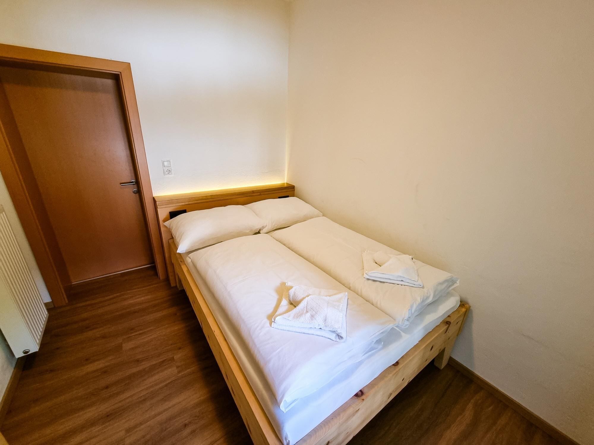 Un lit simple avec du linge de lit blanc et une tête de lit en bois, dans une pièce dotée d’un plancher en bois et d’une porte.