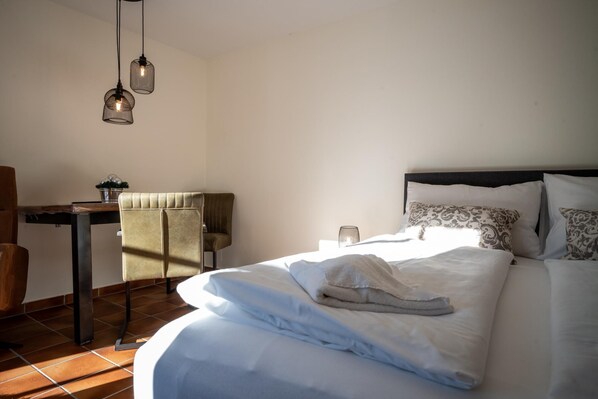 A neatly made bed with white linens, a headboard, and patterned pillows. There is a small table with a plant and a lamp. The room has a tiled floor and a chair.