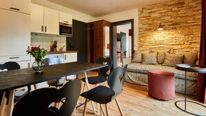 A modern kitchen with white cabinets, a dark countertop, and a dining area with black chairs. A red ottoman and a patterned sofa are visible in the living area. - EVI Apartments - Evi (Kaprun)